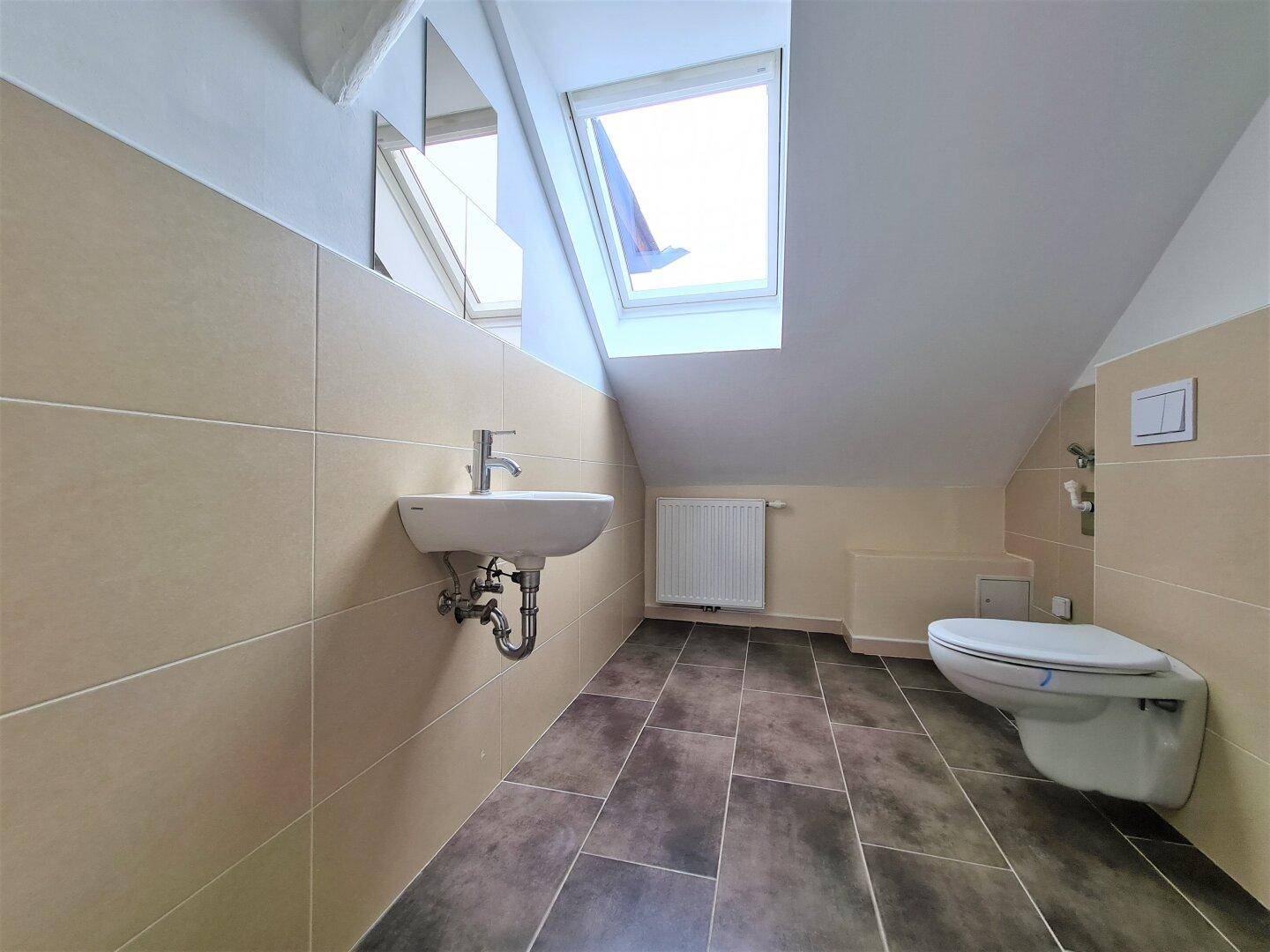 Kleines WC im Dachgeschoss mit Wand-WC, Waschbecken und einem Dachfenster für Tageslicht.