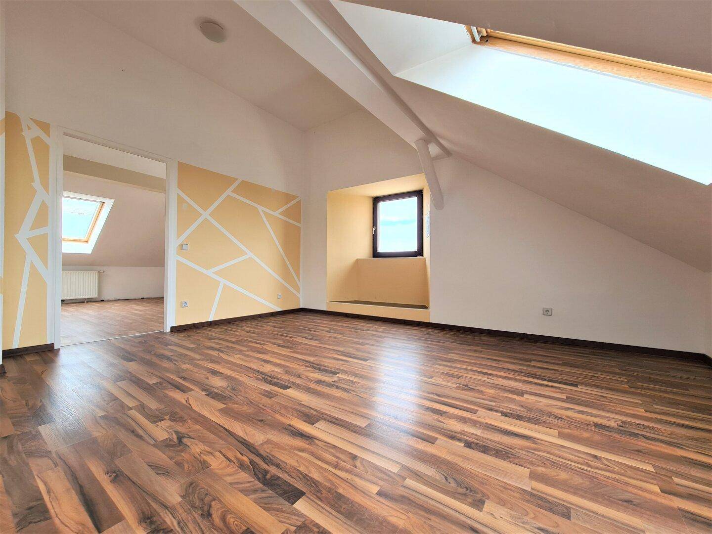 Geräumiger Raum mit Laminatboden, Dachschrägen und einem Dachfenster, teilweise mit geometrischer Wandgestaltung.