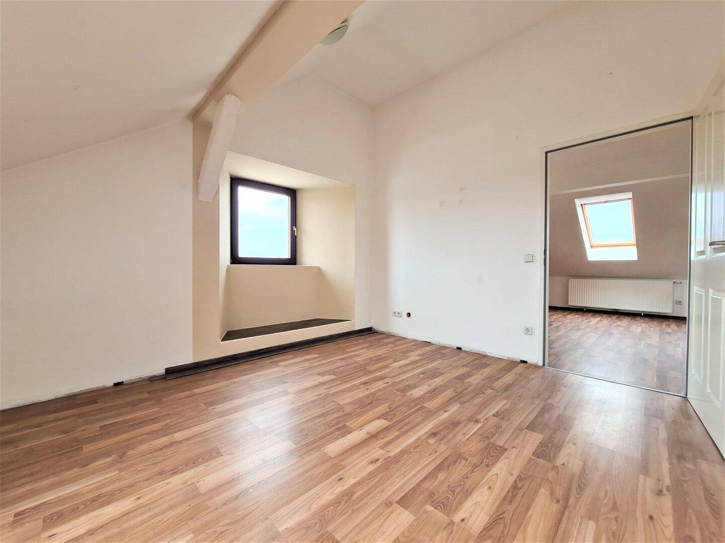 Geräumiger Dachbodenraum mit Laminatboden, schrägen Decken und einem Fenster mit Ausblick.