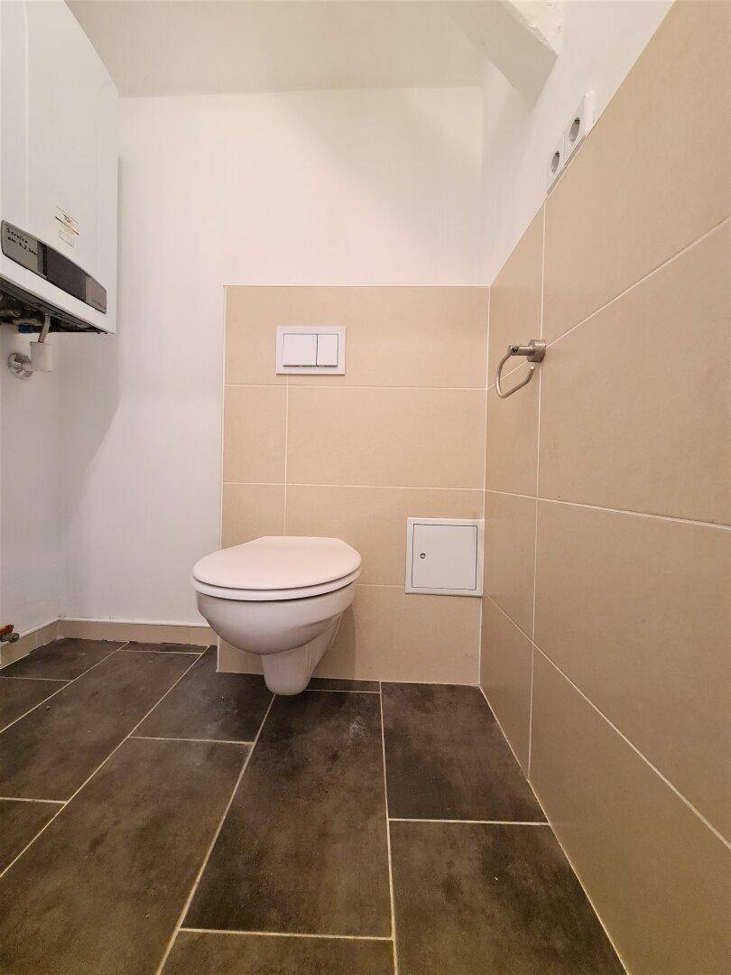 Kleines WC mit wandhängender Toilette, Fliesenboden und einem Warmwasserbereiter an der Wand.