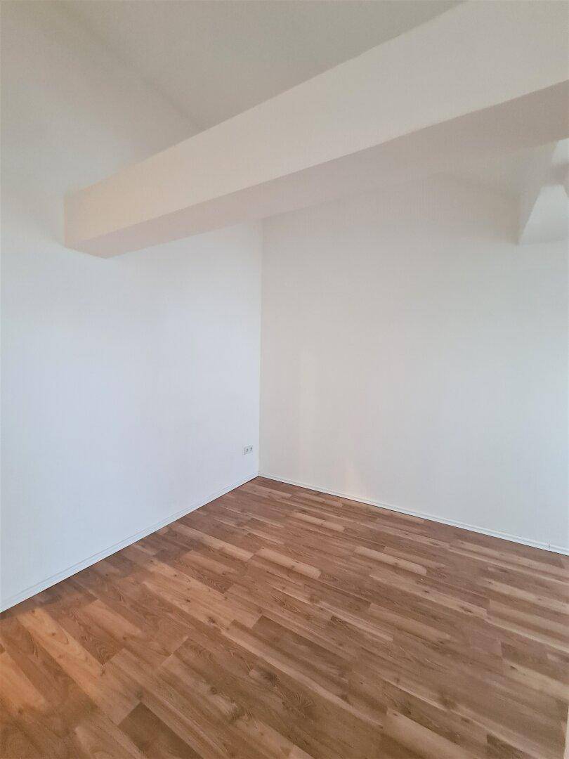 Leerer Raum mit Laminatboden und weißen Wänden, akzentuiert durch einen sichtbaren Deckenbalken.