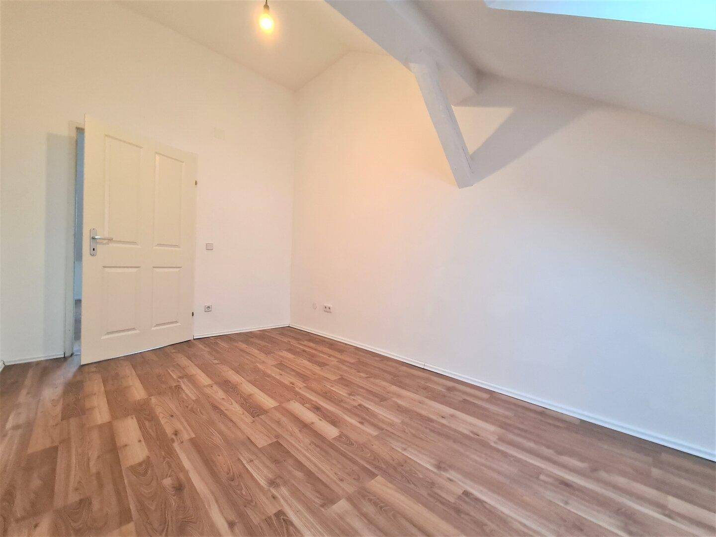 Heller Dachbodenraum mit Laminatboden, schrägen Decken und einer Tür zu einem weiteren Bereich.