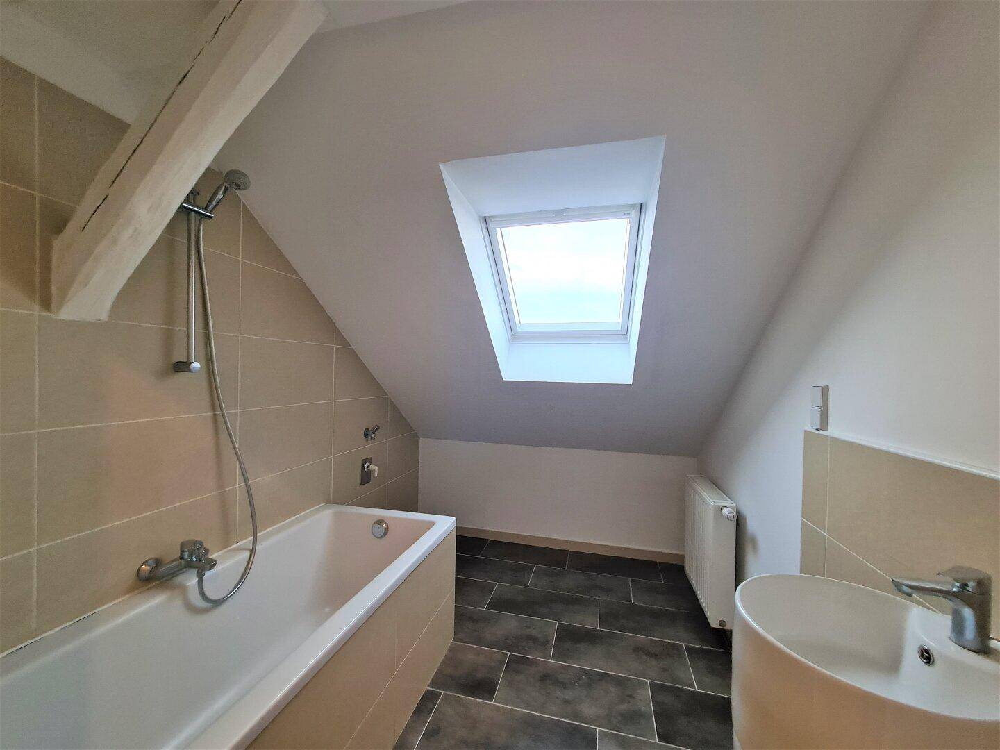 Badezimmer unter Dachschräge mit Badewanne, Waschbecken, Dachfenster und Fliesenboden.