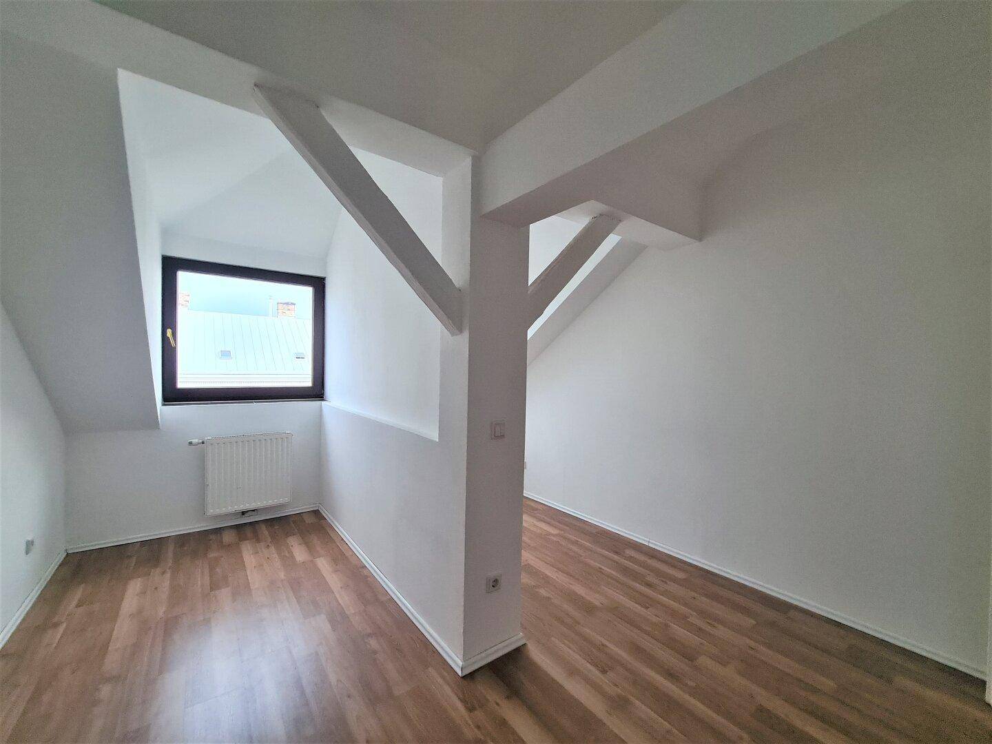 Heller Raum mit Laminatboden, Dachschrägen, einem Fenster und markanten sichtbaren Holzbalken.
