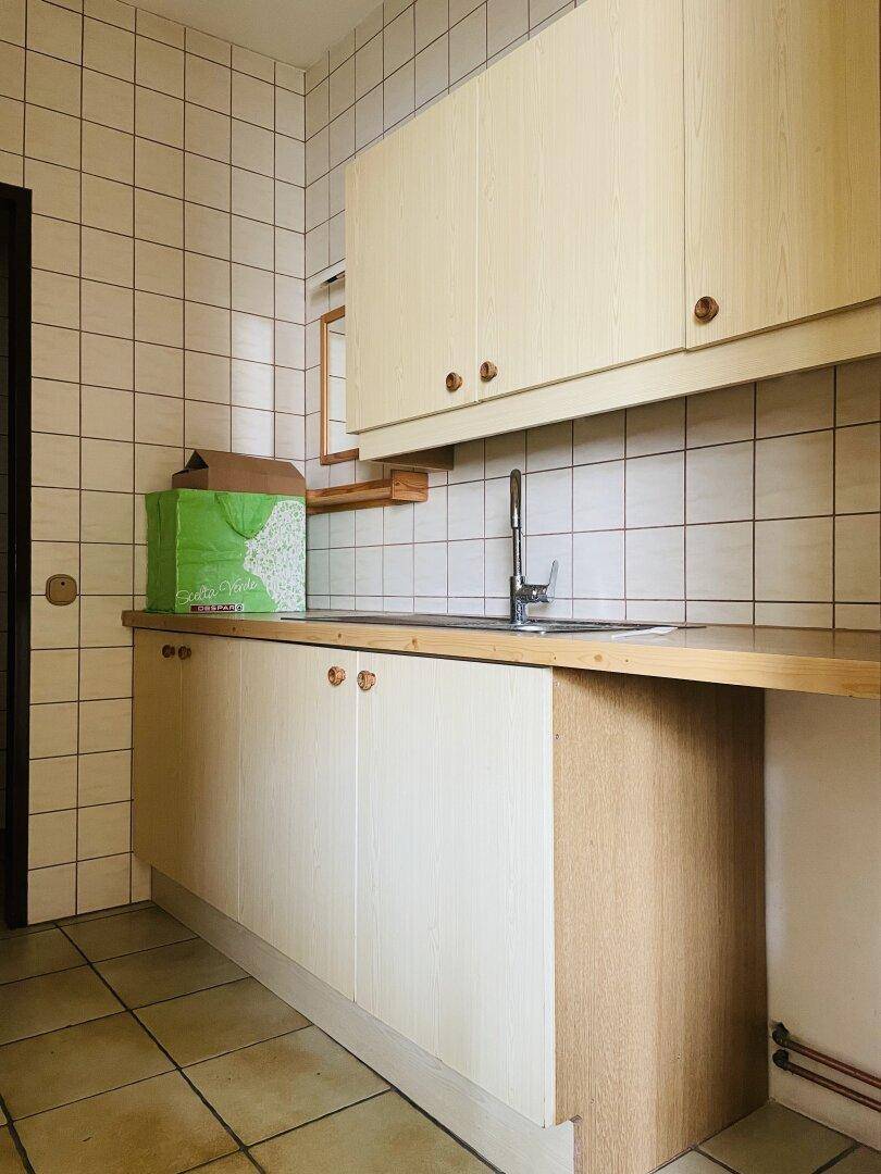 Kleine Küche mit hellen Holzschränken, Arbeitsplatte und Spüle, umgeben von weißen Wandfliesen.