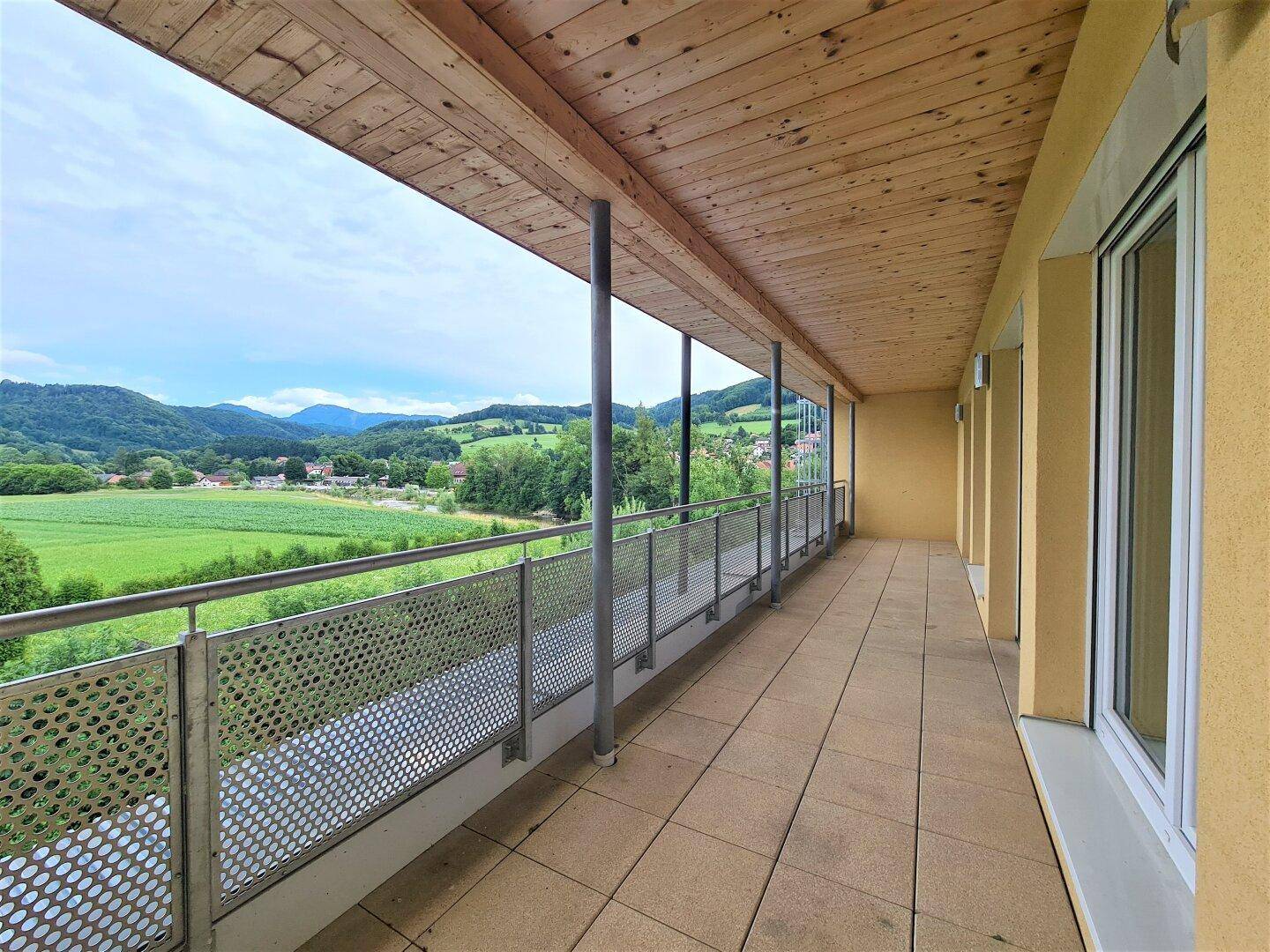 Langer überdachter Balkon mit Fliesenboden und weitem Blick über die grüne Landschaft.