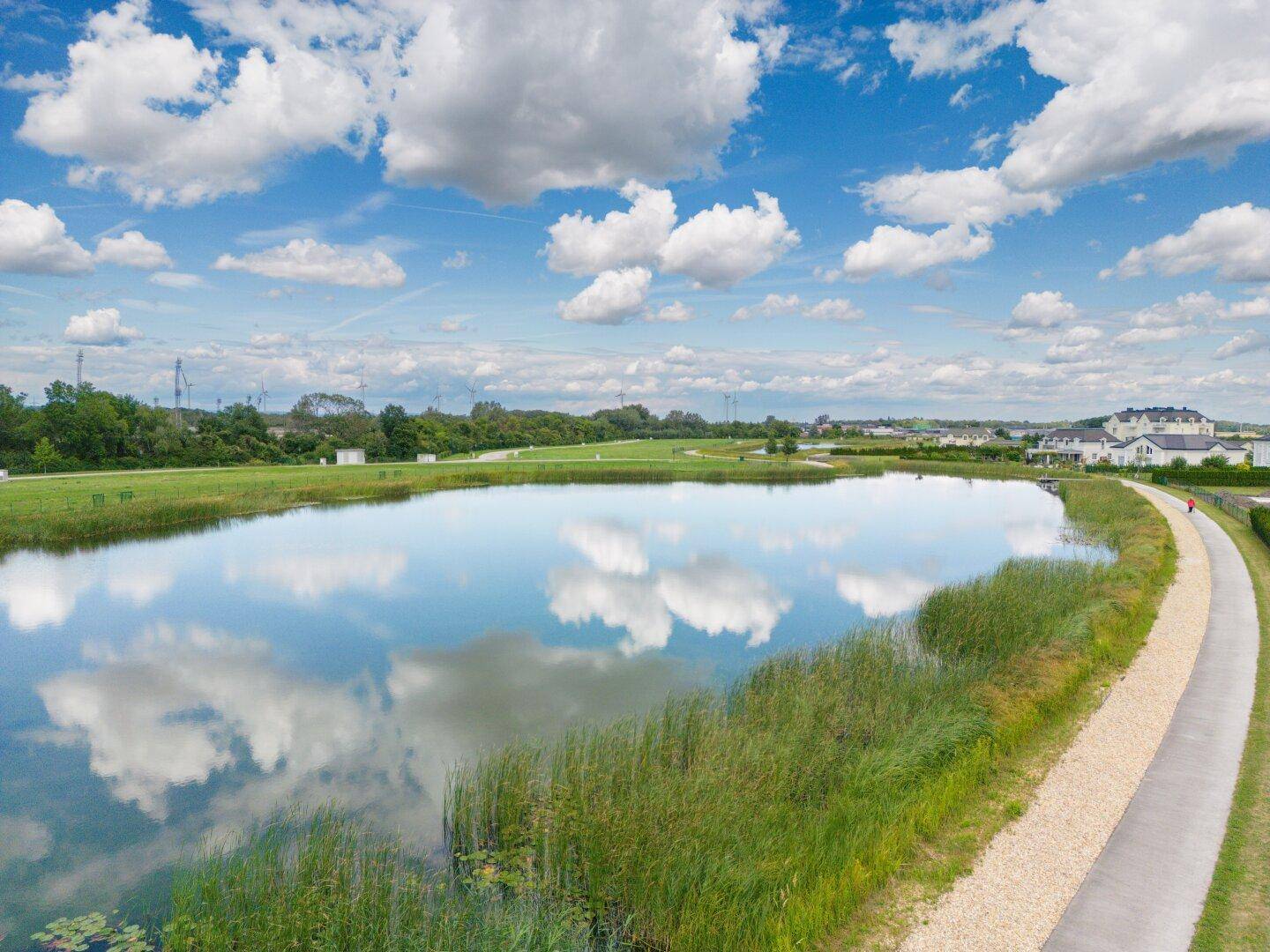 Weitläufige Luftaufnahme einer Wohnsiedlung mit großem Teich, umgeben von grünen Feldern und Windrädern am Horizont.
