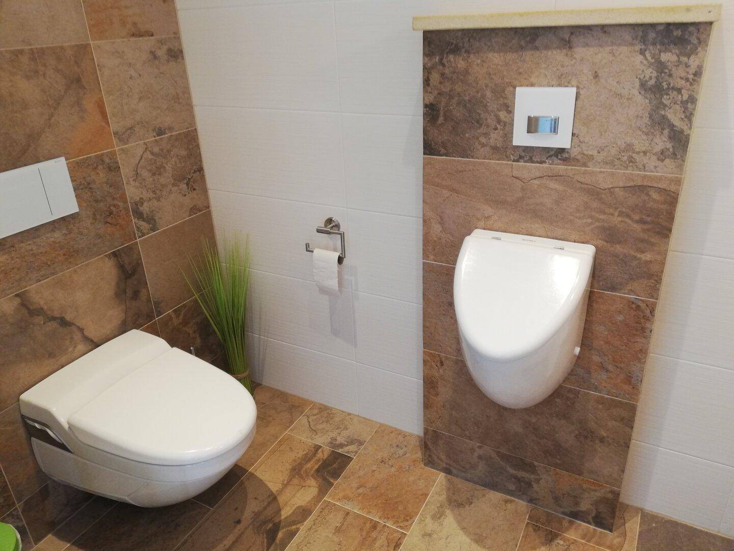 Modernes WC mit wandhängender Toilette, Urinal und stilvollen Fliesen an Wänden und Boden.