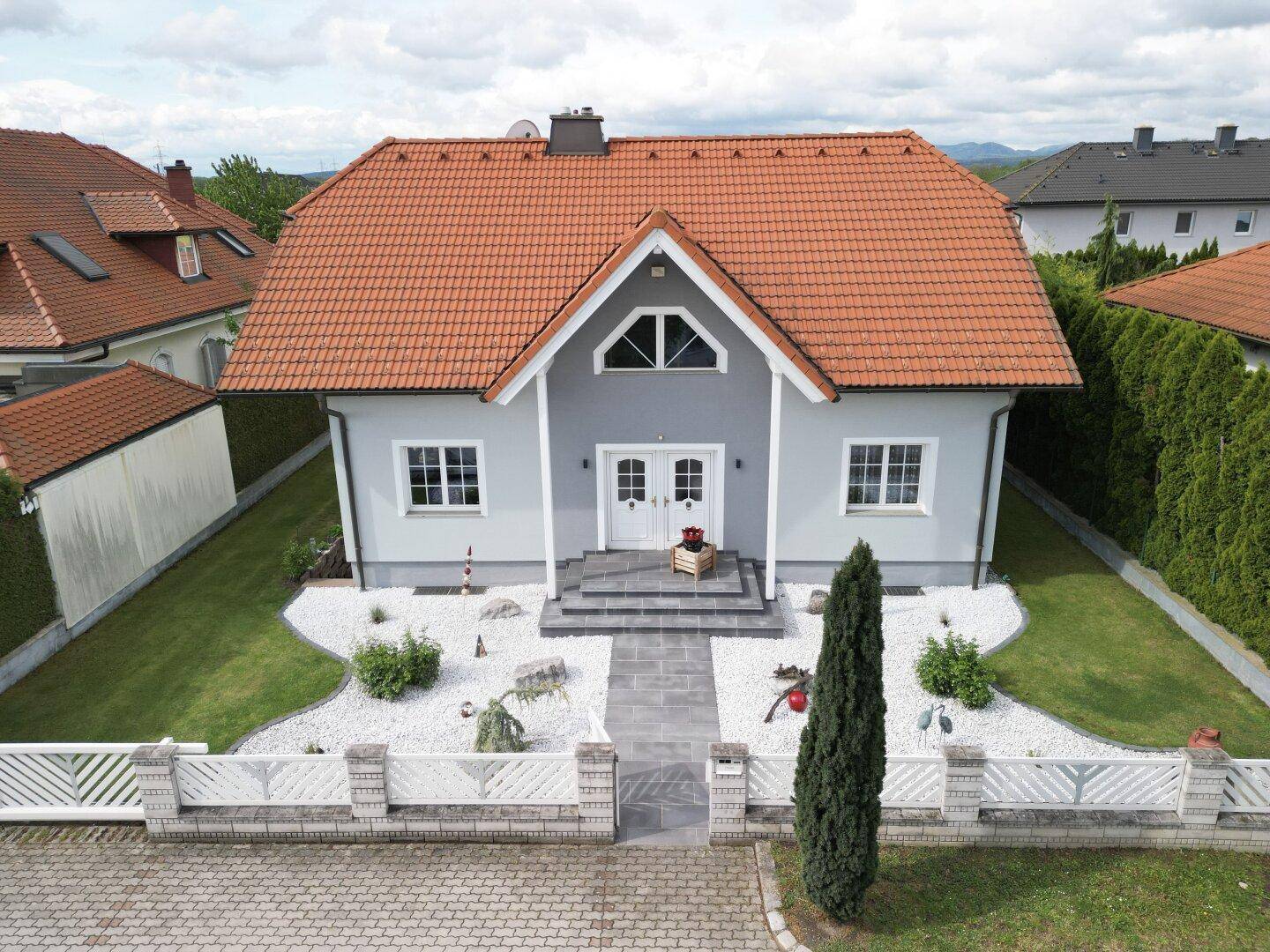 Modernes Einfamilienhaus mit hellgrauer Fassade, rotem Ziegeldach und einem schön angelegten Vorgarten.