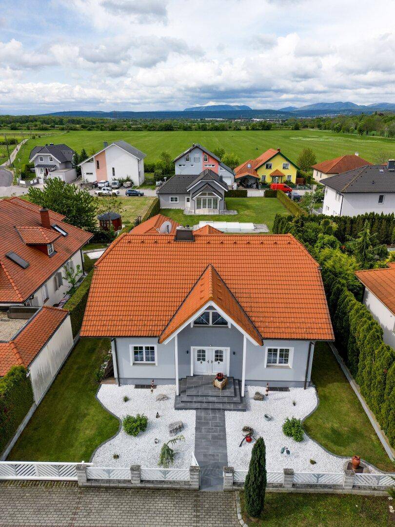 Luftaufnahme eines gepflegten Einfamilienhauses mit Vorgarten, umgeben von grünen Feldern und Bergen im Hintergrund.