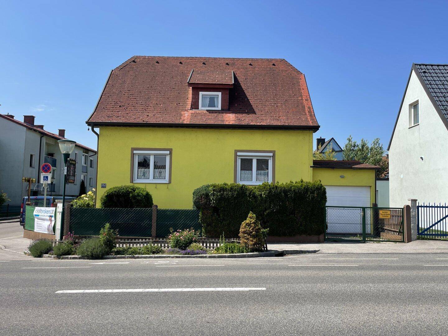 Vorderansicht eines gelben Hauses mit rotem Dach, umgeben von gepflegten Hecken und einem Garagentor.
