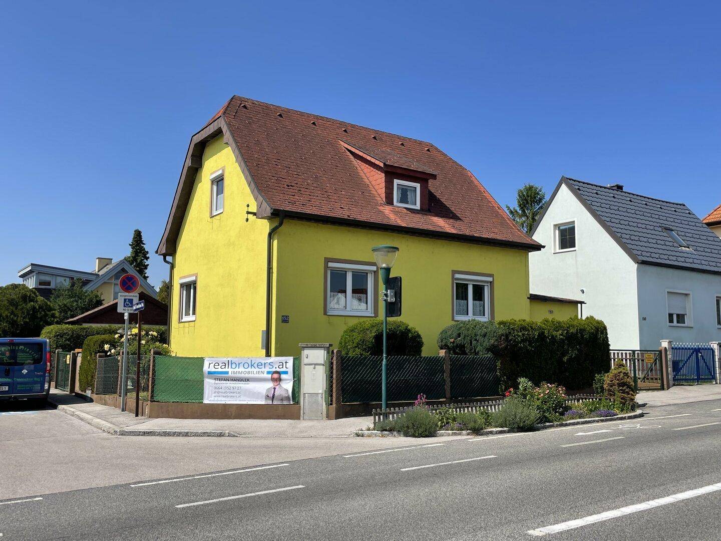 Gelbes Einfamilienhaus an einer Straßenecke mit rotem Dach und gepflegter Hecke unter blauem Himmel.