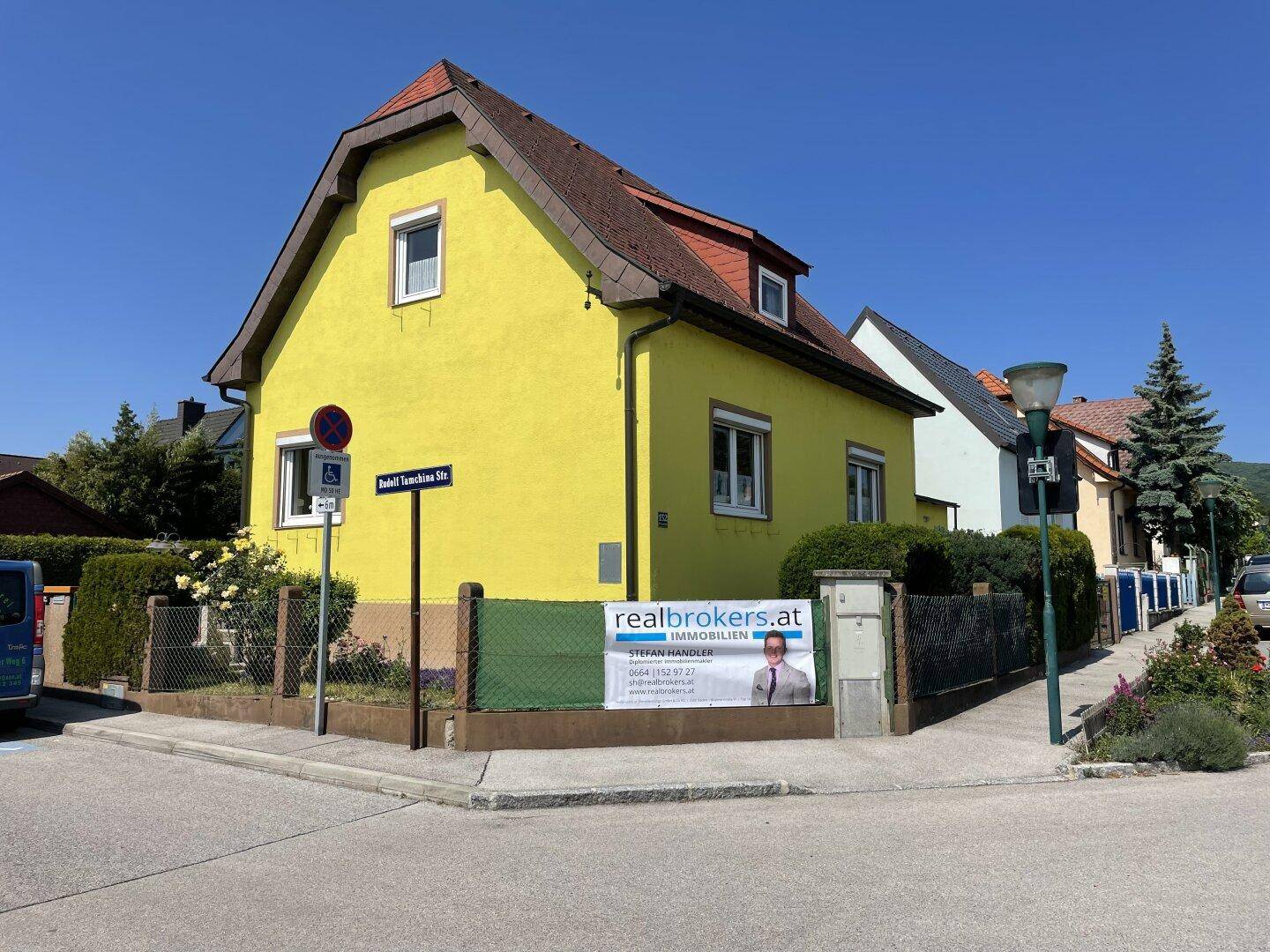 Gelbes Einfamilienhaus mit rotem Ziegeldach an einer Straßenecke, umgeben von Hecken und einem Zaun.