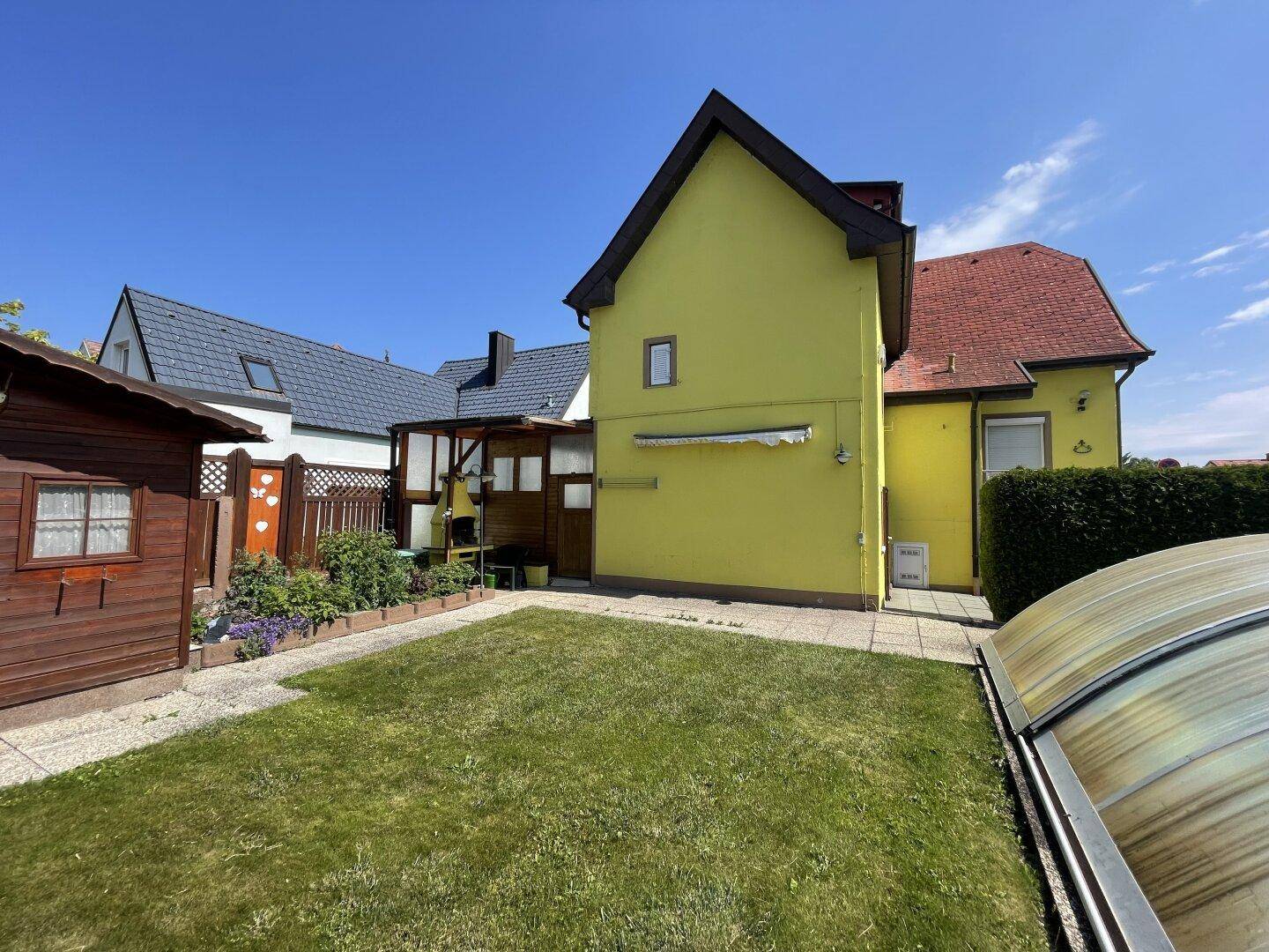 Hinterhof mit Rasenfläche, Terrasse, Gartenhaus und einem überdachten Grillbereich neben dem gelben Haus.
