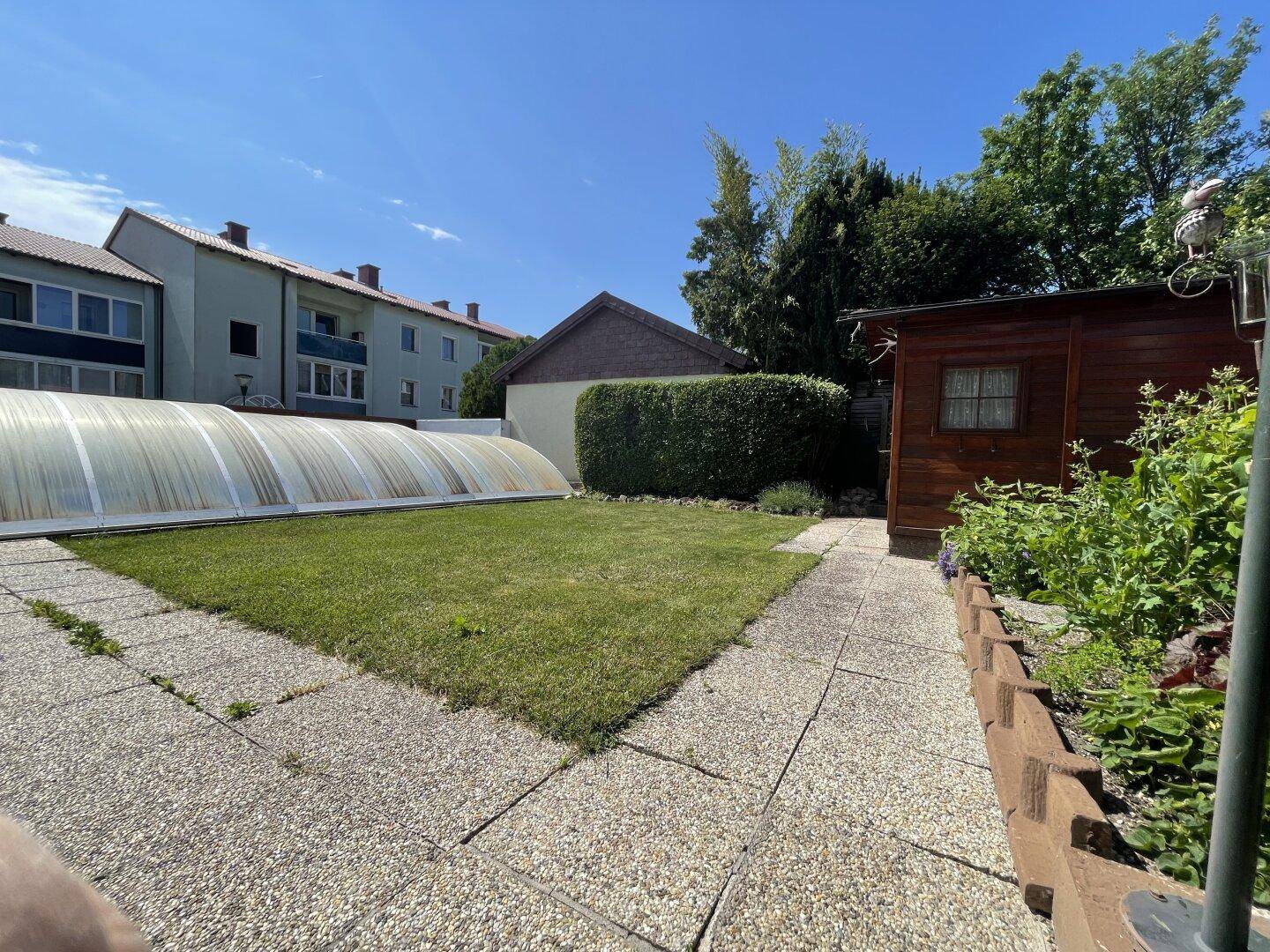 Gepflegter Garten mit Rasen, Steinplattenweg, Gartenhaus und einer Poolabdeckung im Sonnenlicht.