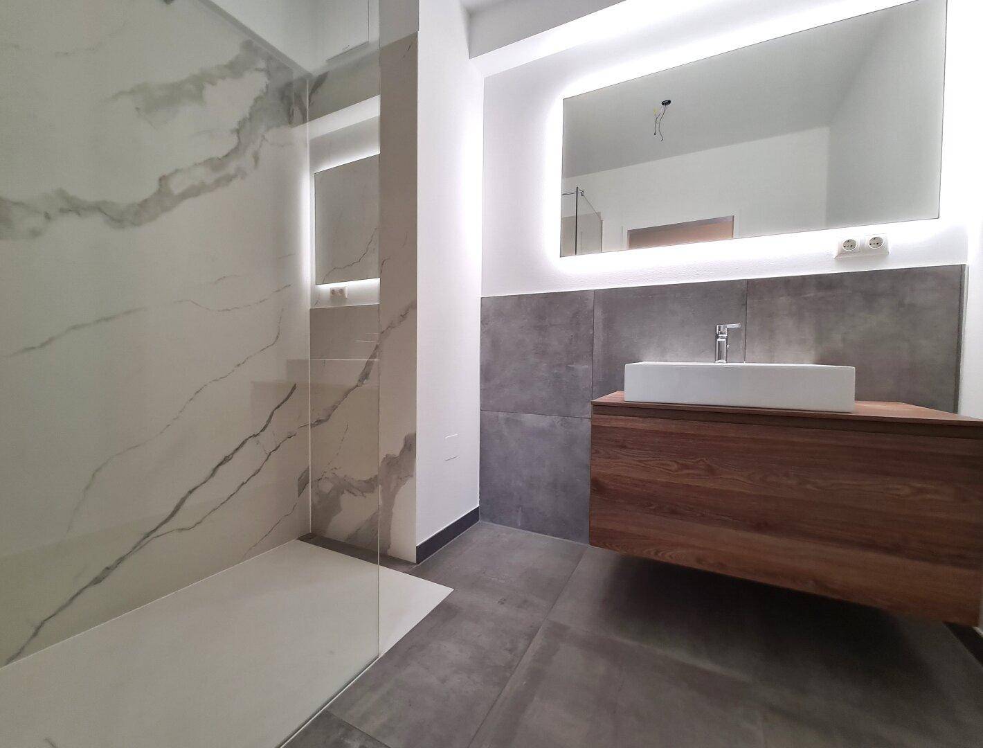 Modernes Badezimmer mit großer ebenerdiger Dusche, Holzwaschtisch und beleuchtetem Spiegel.
