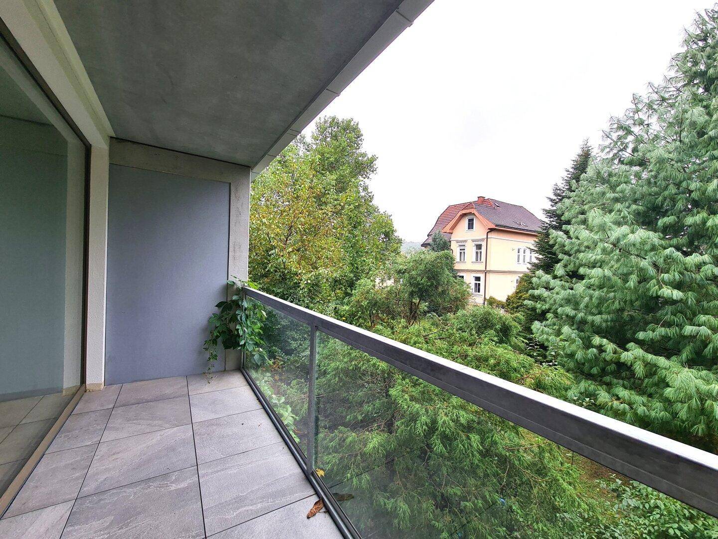 Moderner Balkon mit Fliesenboden, Glasgeländer und weitem Blick ins Grüne mit einem Haus im Hintergrund.