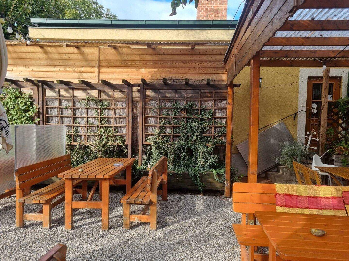 Rustikaler Außenbereich mit Holzbänken und Tischen, umgeben von begrünten Rankgittern.