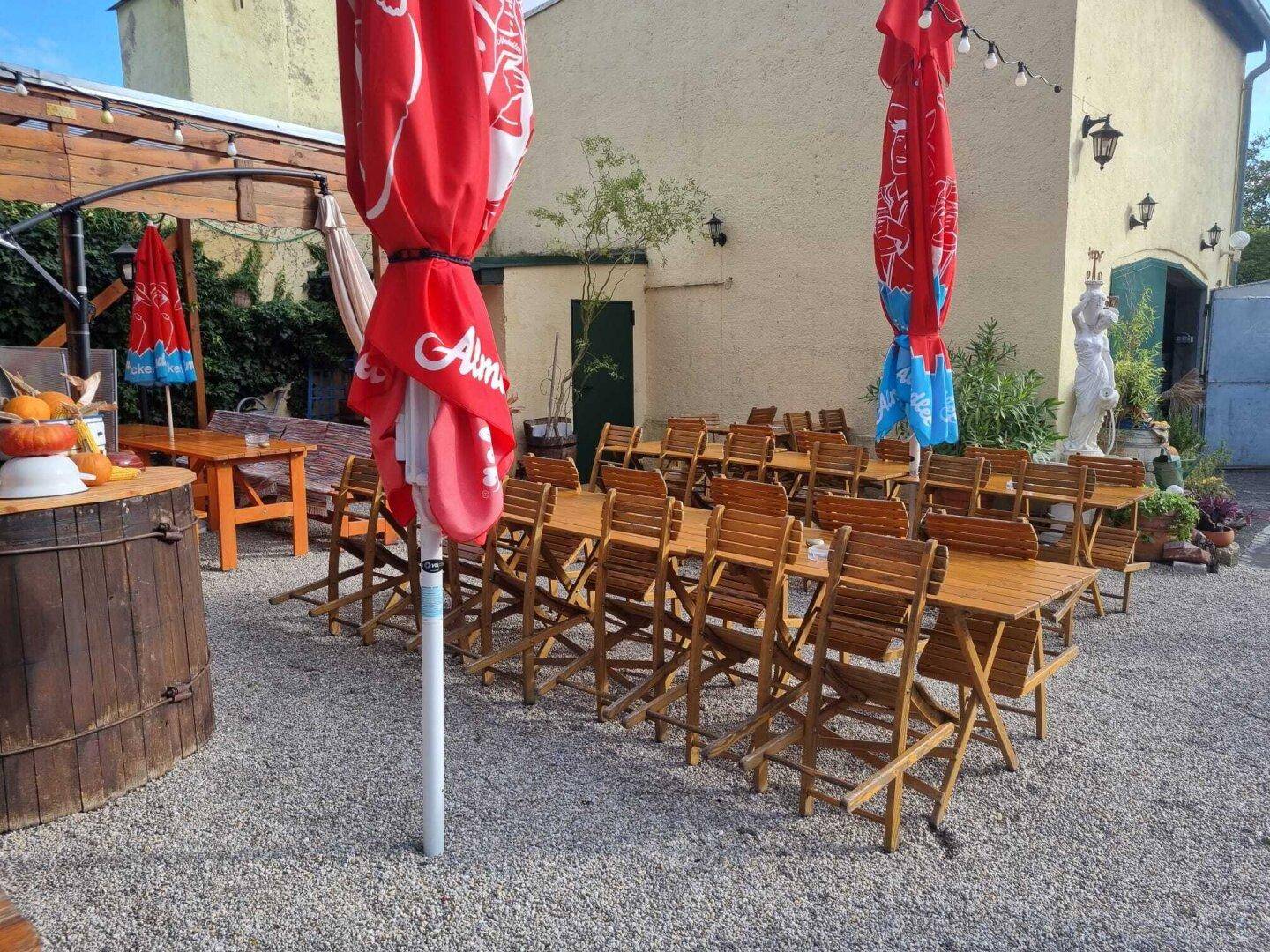 Geräumiger Außenbereich mit vielen Holztischen und roten Sonnenschirmen vor einer hellen Gebäudefassade.