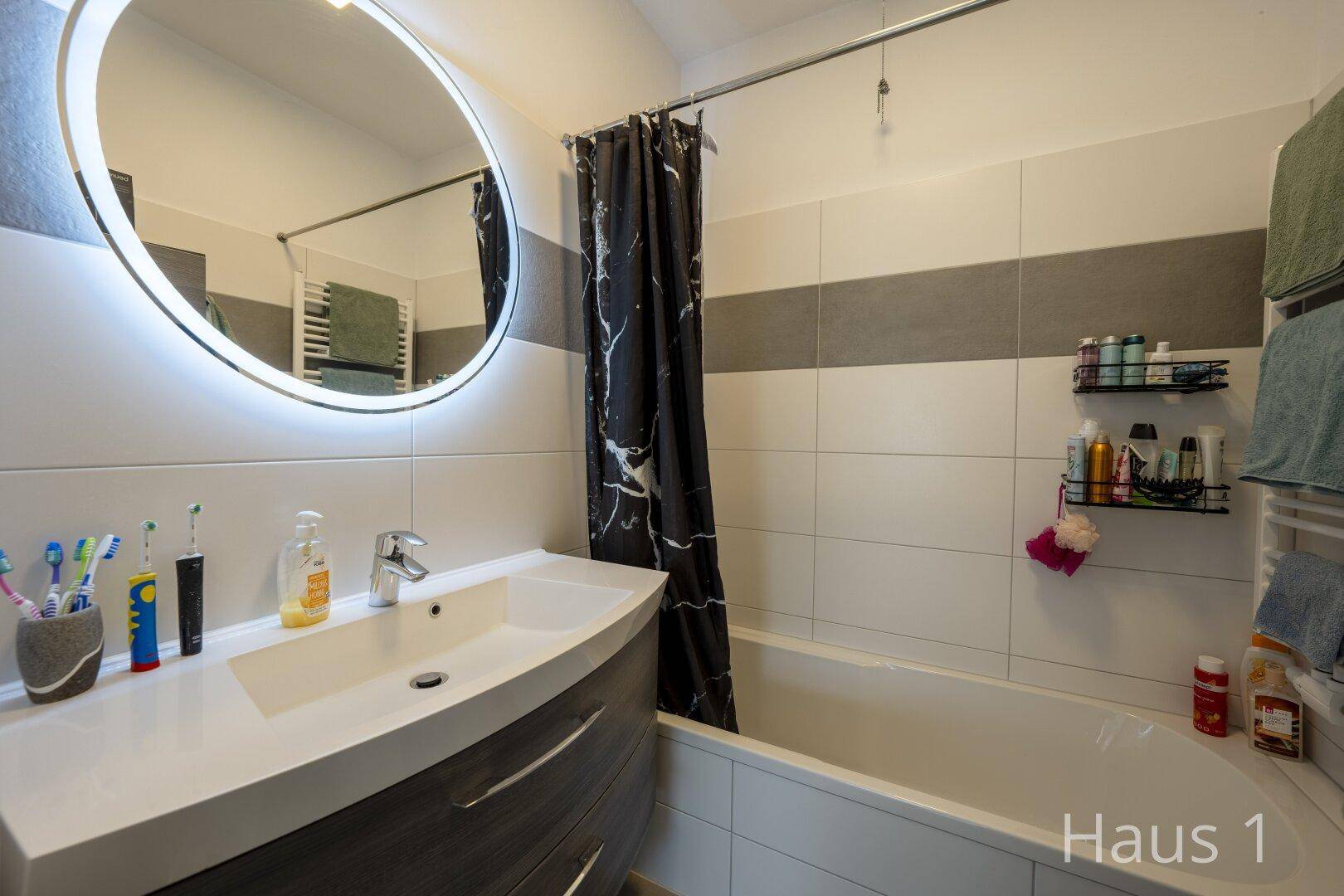 Modernes Badezimmer mit Badewanne, beleuchtetem Spiegel und grauen sowie weißen Fliesen.