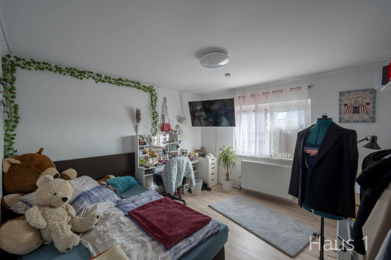 Helles Schlafzimmer mit Bett, Schreibtisch und Fenster, dekoriert mit Pflanzen und Bildern.