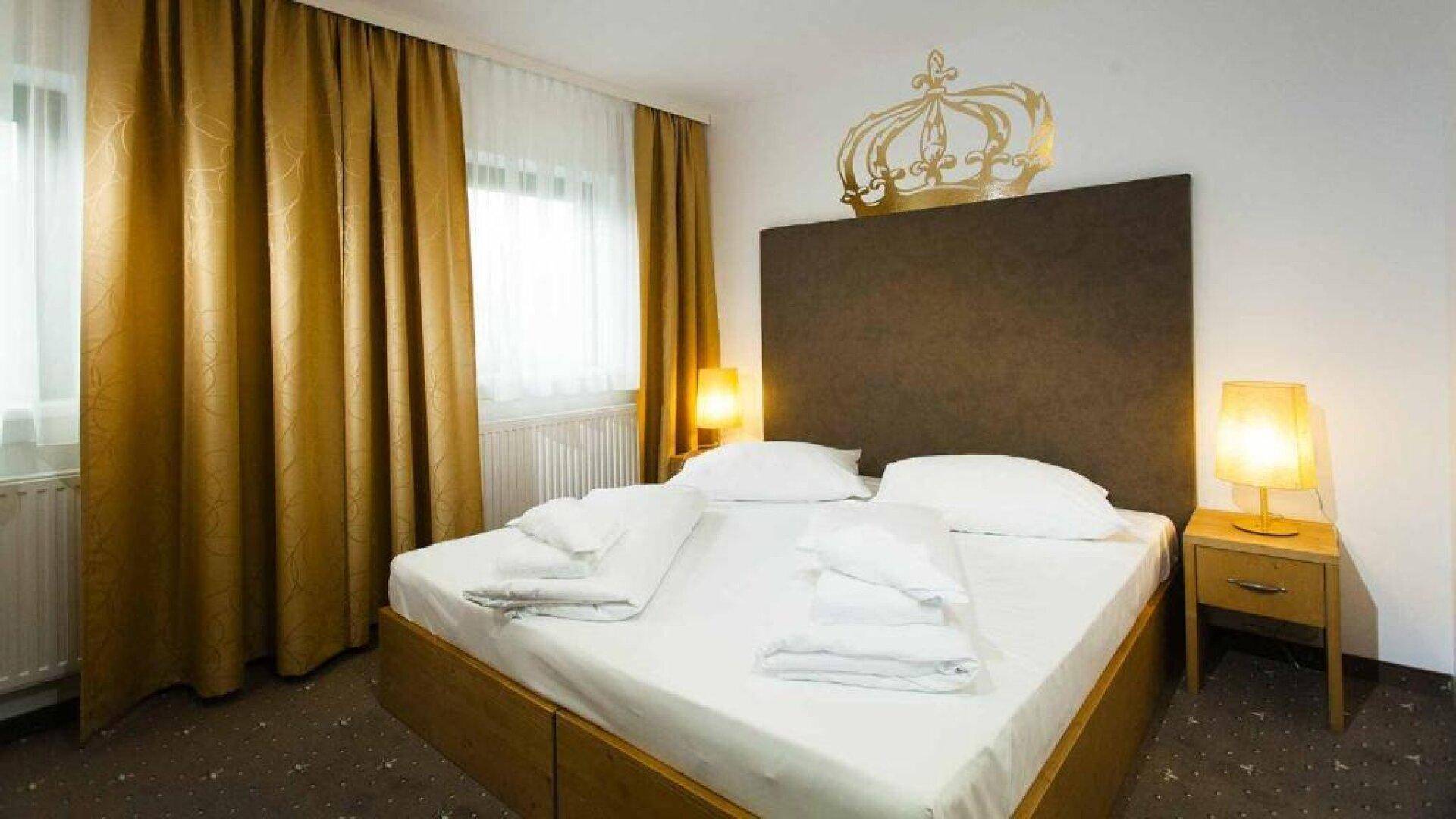 Schlafzimmer mit Doppelbett, Nachttischen, goldenen Vorhängen und einer Kronendekoration an der Wand.