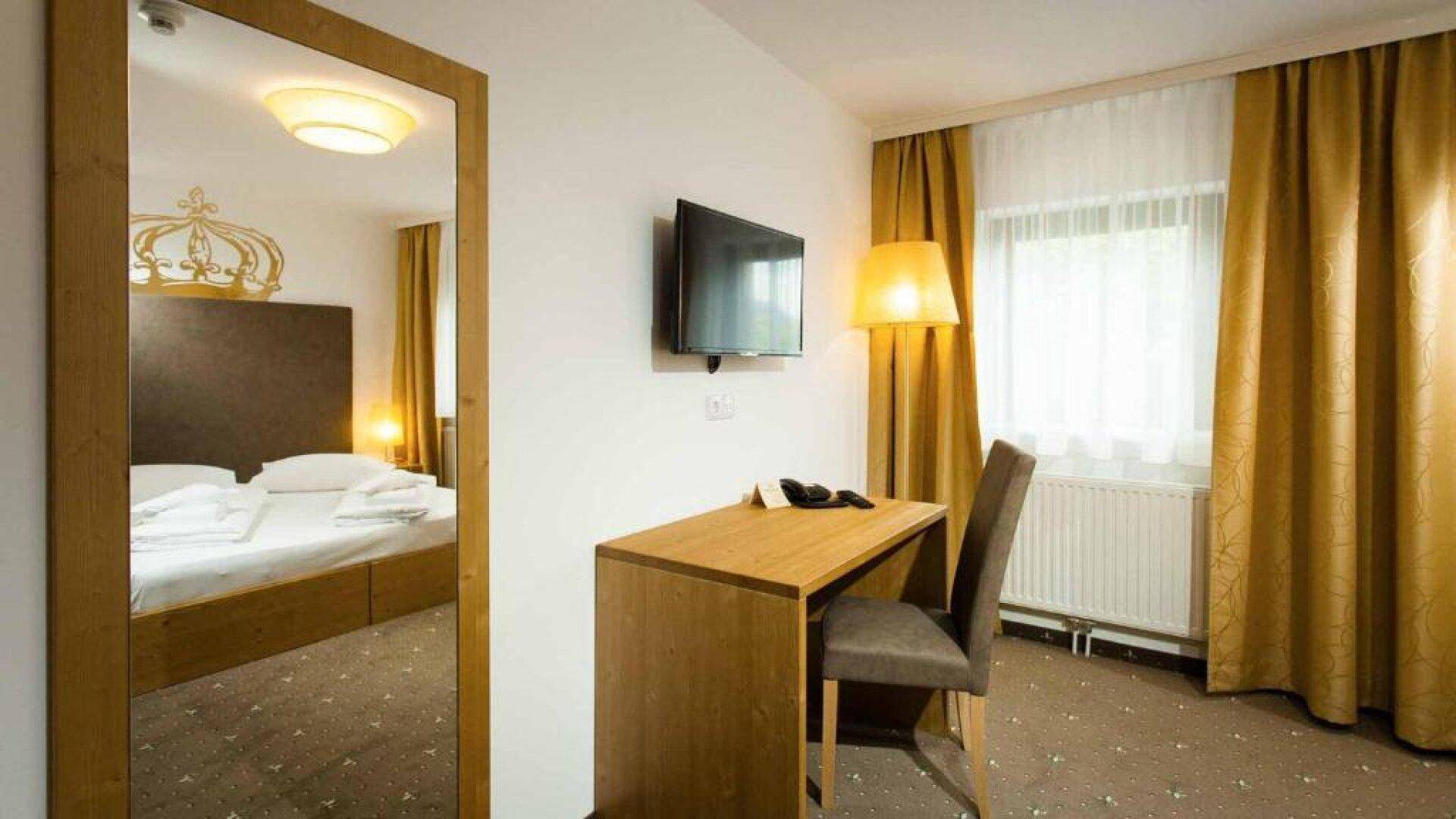 Schlafzimmer mit Schreibtisch, Stuhl und Wandfernseher, ein großer Spiegel reflektiert das Bett.