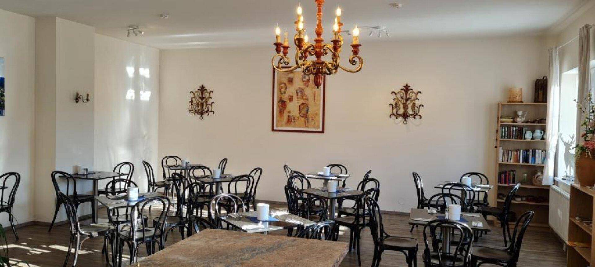 Heller Speisesaal mit vielen kleinen Tischen, Stühlen, einem Kronleuchter und einem Bücherregal.