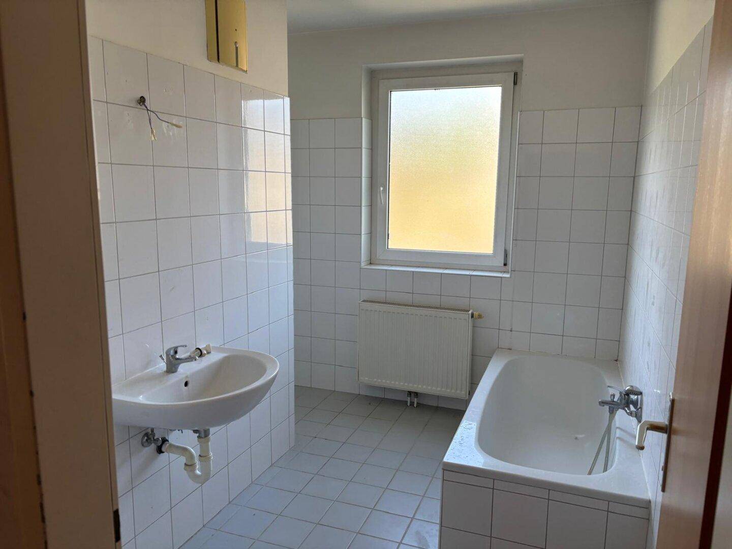 Badezimmer mit weißen Fliesen, Waschbecken, Badewanne und einem Fenster.