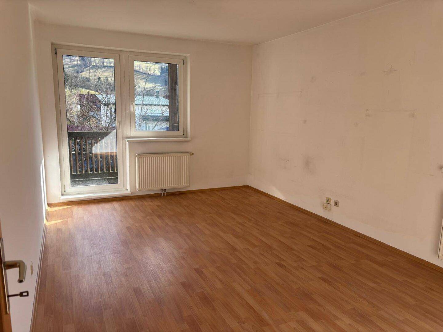 Leerer Raum mit Laminatboden und Fensterfront, die einen Zugang zum Balkon bietet.