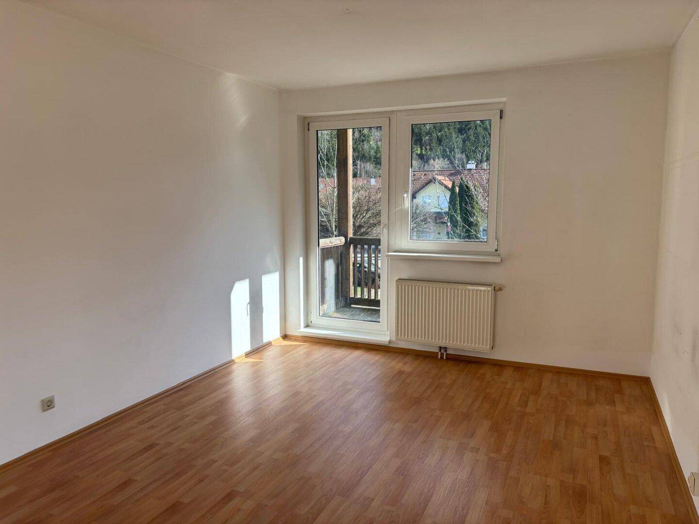 Heller, leerer Raum mit Laminatboden, Heizkörper und Zugang zu einem kleinen Balkon.
