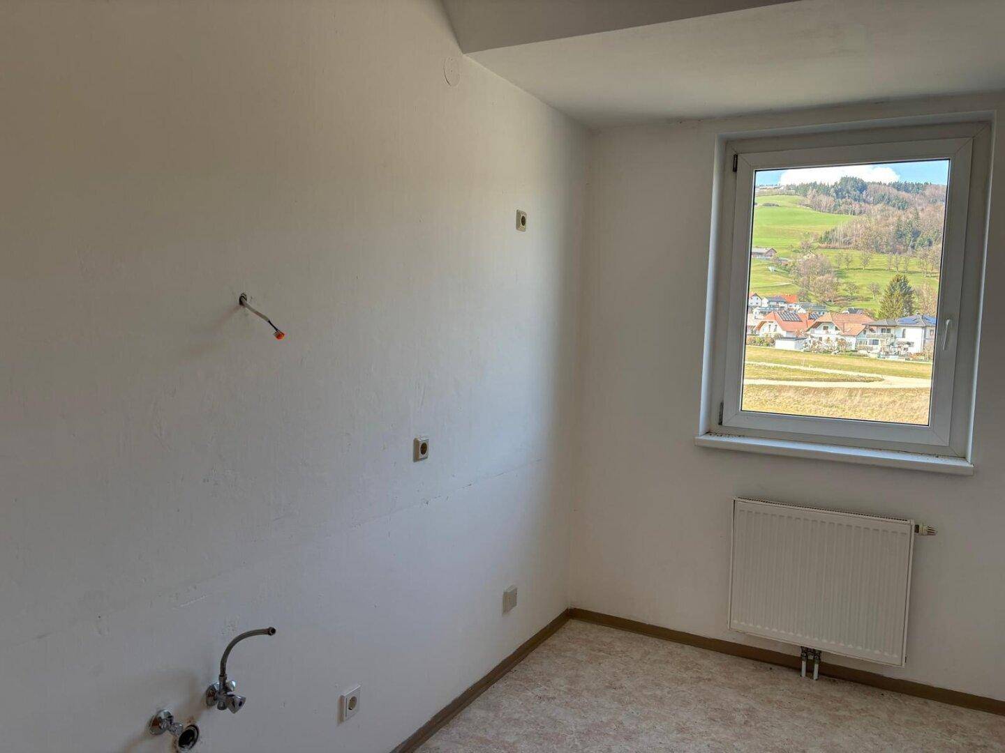 Leerer Küchenraum mit Anschlüssen für Geräte und einem Fenster mit Blick ins Grüne.
