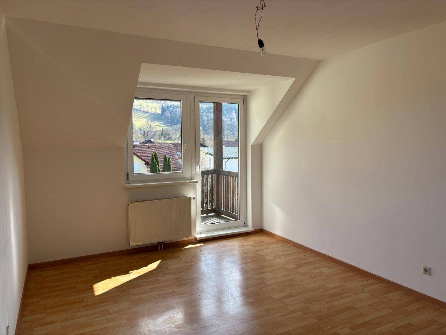 Heller Raum im Dachgeschoss mit Holzboden, schrägen Wänden und Zugang zu einem kleinen Balkon.