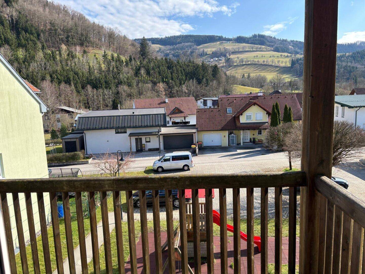 Blick vom Balkon auf ein Dorf mit Häusern, einem Spielplatz und bewaldeten Hügeln im Hintergrund.