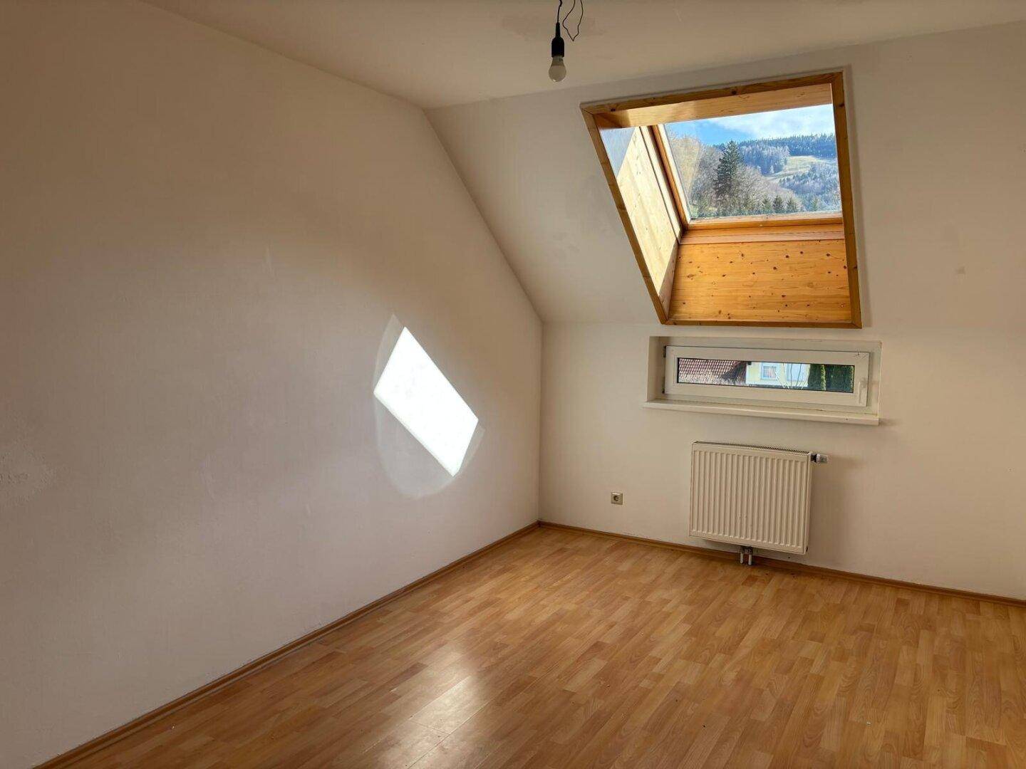 Leeres Zimmer mit Laminatboden, weißen Wänden und einem Dachfenster, das viel Tageslicht hereinlässt.