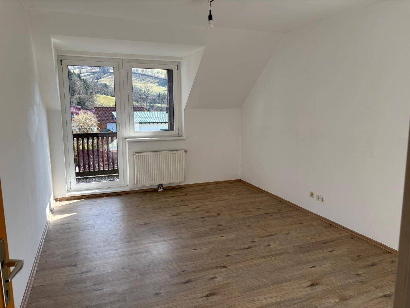 Leeres Zimmer mit Laminatboden, weißen Wänden und Balkontür, die einen Blick auf die Landschaft bietet.