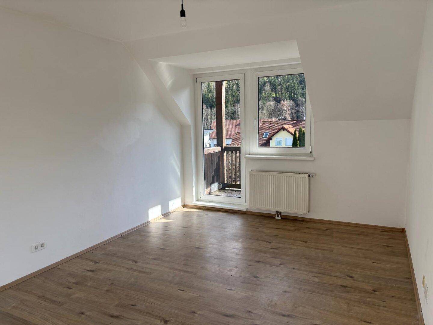 Helles, leeres Zimmer mit Laminatboden, weißen Wänden und Zugang zu einem kleinen Balkon mit Bergblick.