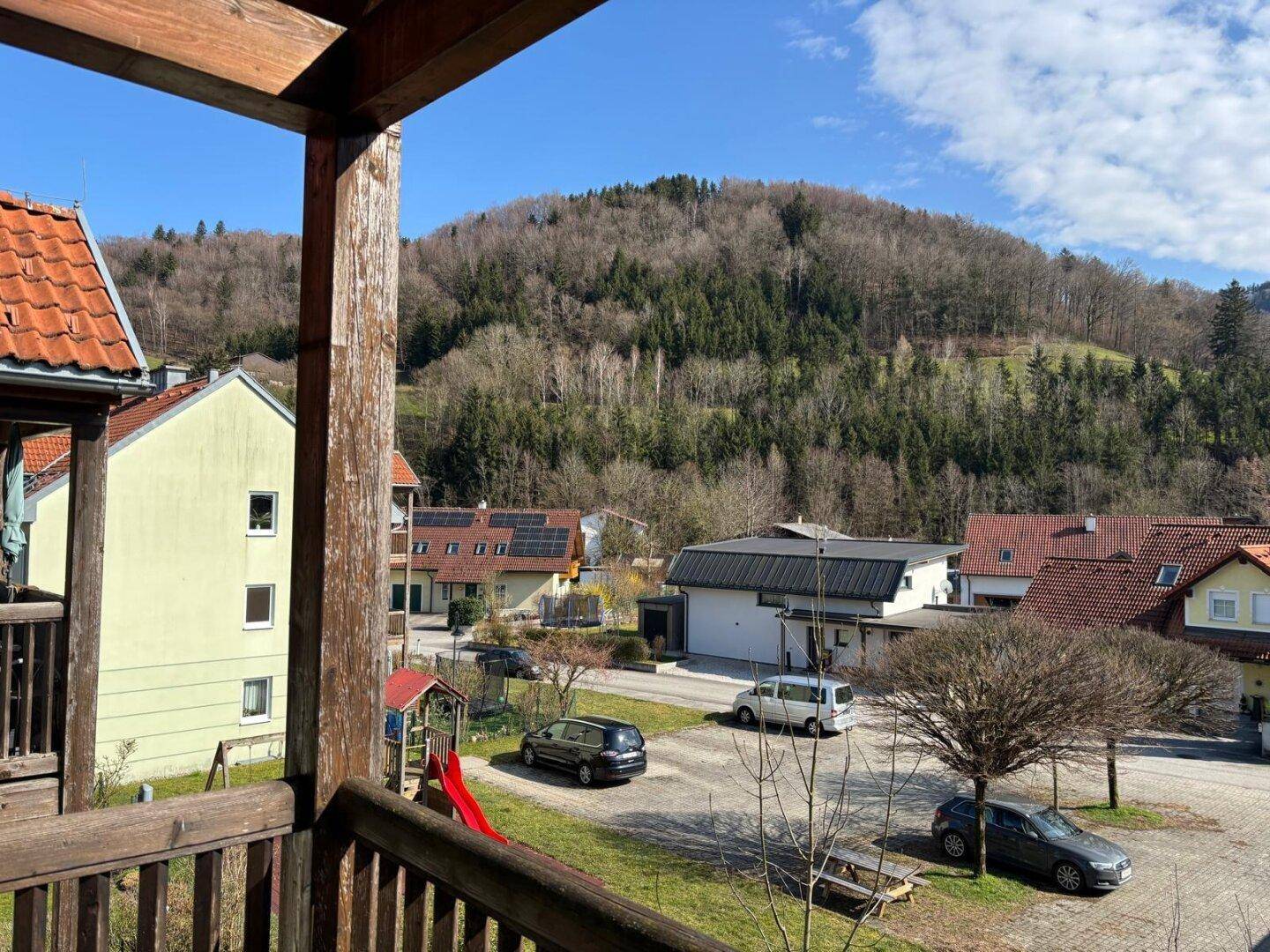 Aussicht von einem Balkon auf eine Wohnsiedlung mit Spielplatz und einem bewaldeten Berg im Hintergrund.