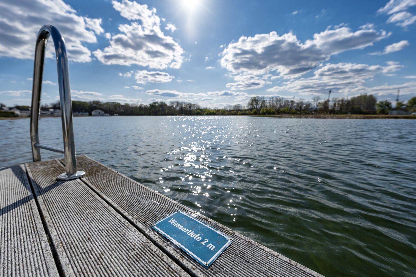 Holzsteg mit Badeleiter und einem Schild für die Wassertiefe von 2 Metern unter strahlendem Sonnenschein.