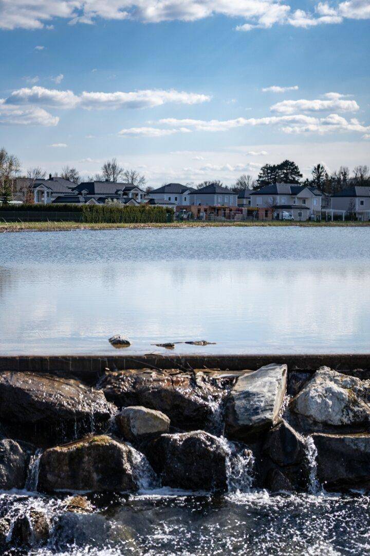 Wasser fließt über Felsen, mit einem See und Häusern im Hintergrund unter blauem Himmel.