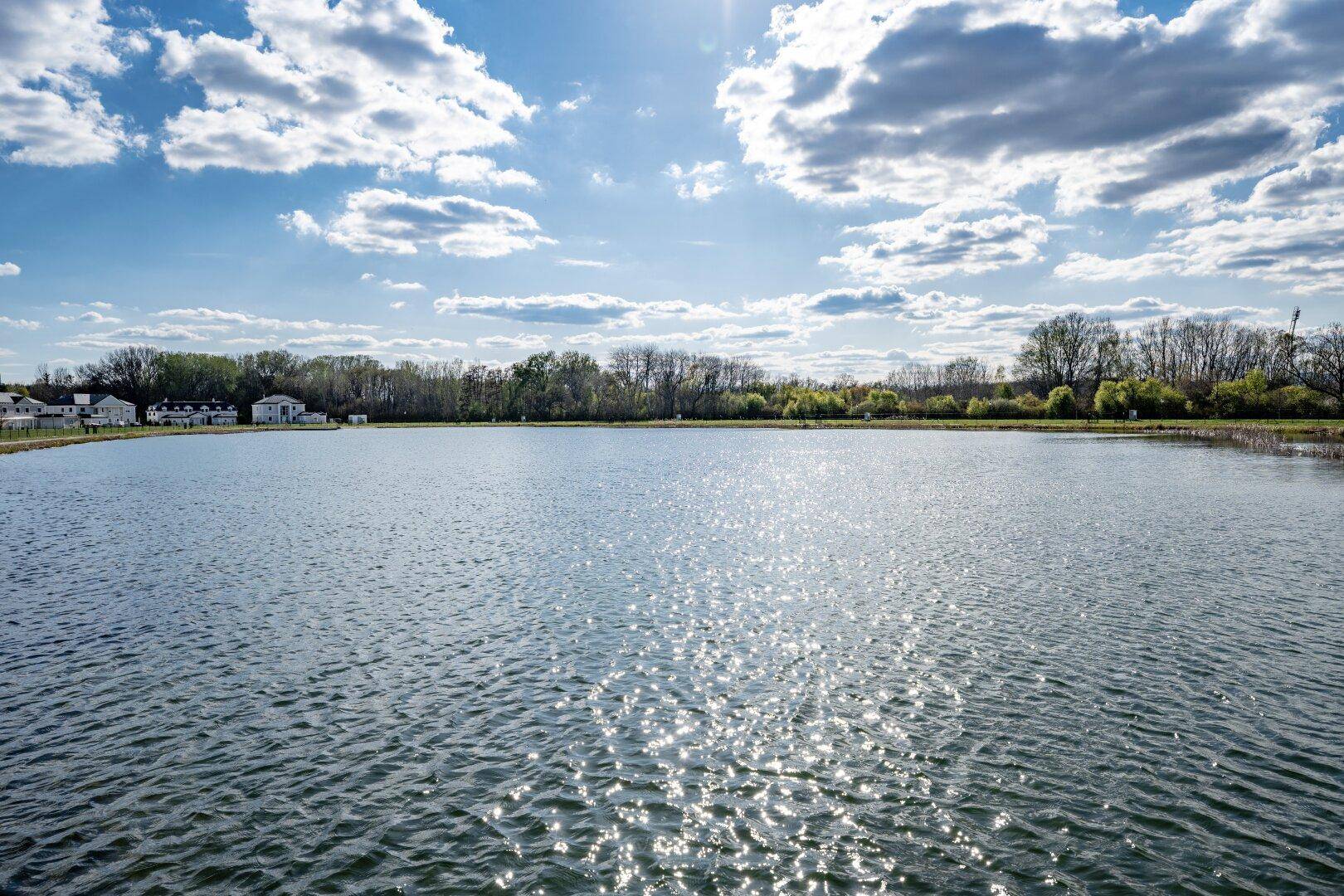 Weitläufiger Blick auf einen See unter sonnigem Himmel mit Wolken und entfernten Häusern.