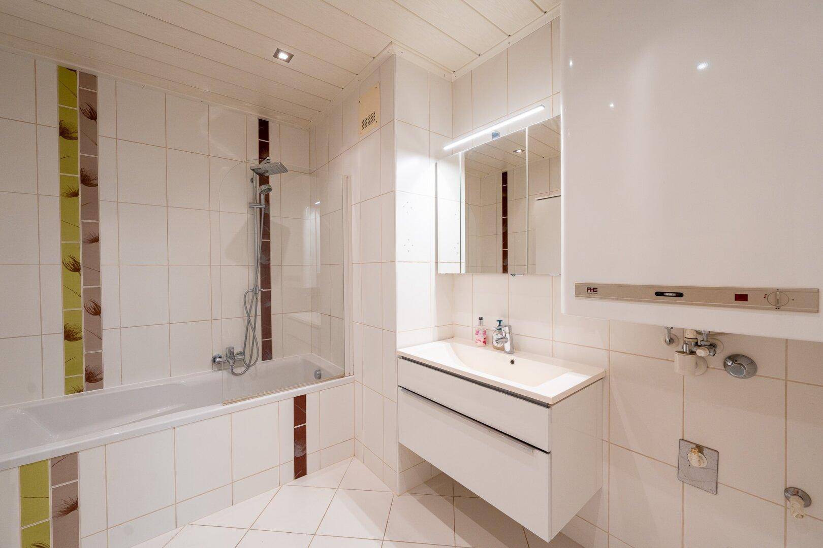Helles Badezimmer mit Badewanne, Dusche und weißen Fliesen, sowie Holzdeckenpaneelen.