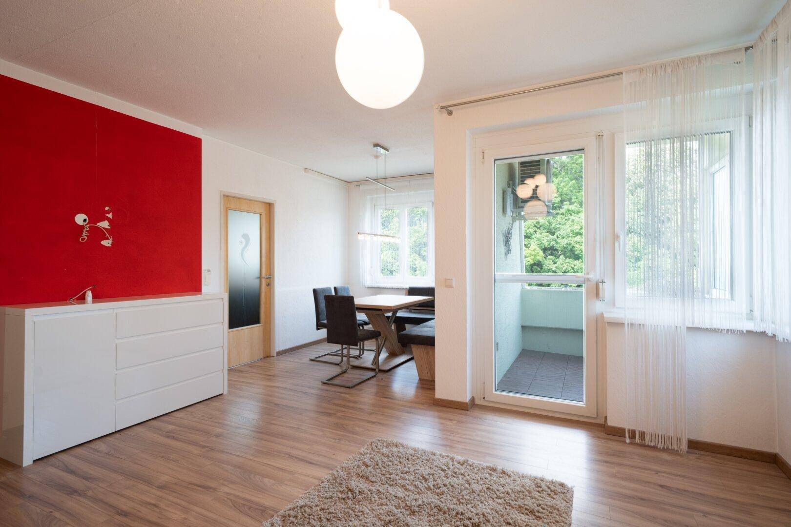 Heller Wohn- und Essbereich mit Holzboden, roter Akzentwand und Zugang zum Balkon.