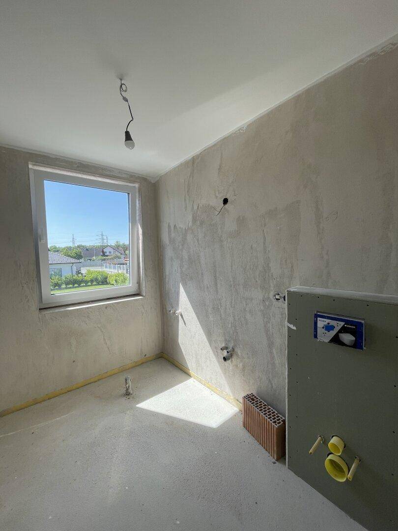 Unfertiges Badezimmer mit Fenster und vorbereiteten Anschlüssen für Sanitäranlagen.