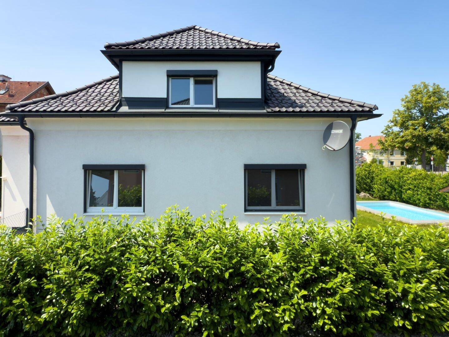 Seitenansicht des Hauses mit Dachgaube, dunklen Fenstern und einer gepflegten Hecke im Vordergrund.