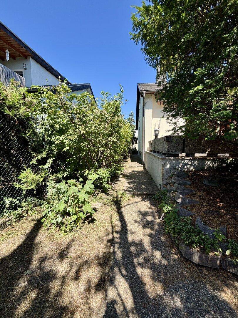 Schmaler Weg zwischen zwei Häusern, umgeben von dichtem Grün und Schatten.