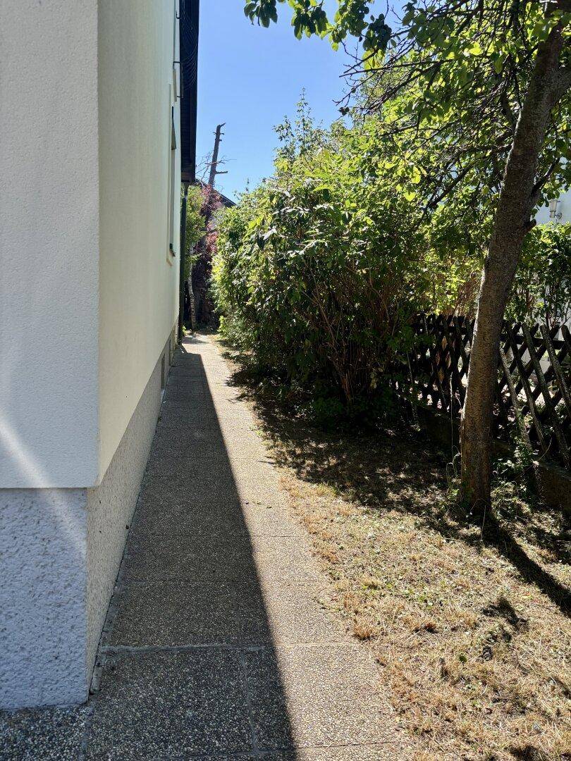 Schmaler gepflasterter Weg entlang der Hausseite, umgeben von Grün und Schatten.
