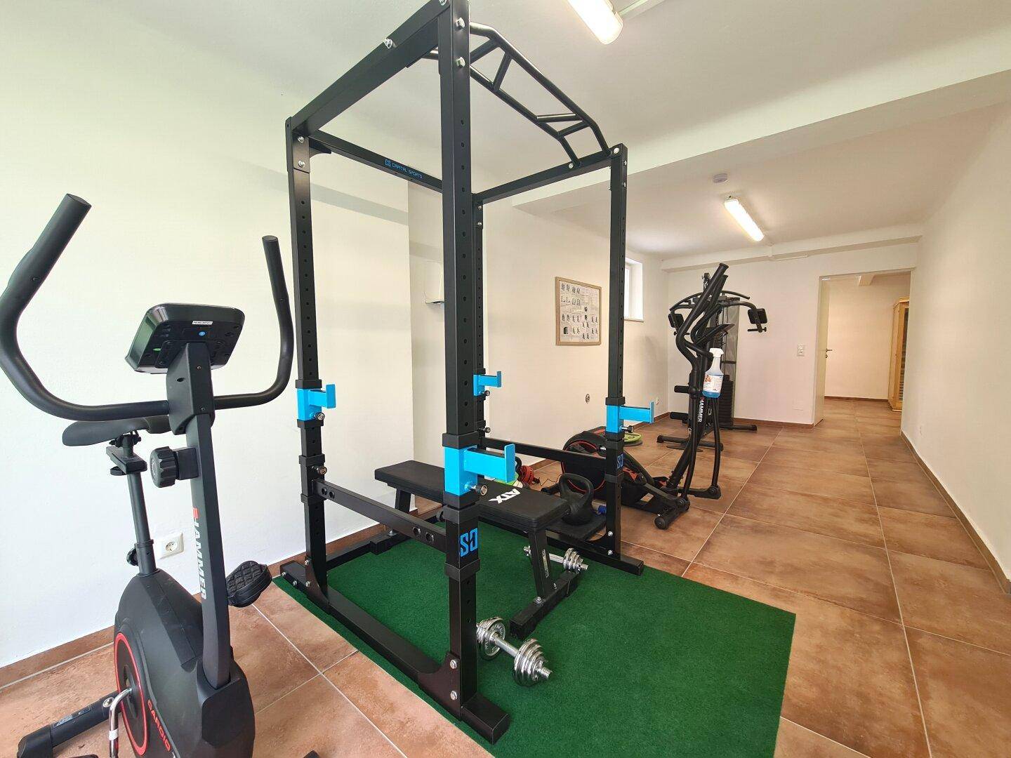 Gut ausgestatteter Fitnessraum mit verschiedenen modernen Trainingsgeräten, Fliesenboden und natürlichem Lichteinfall.