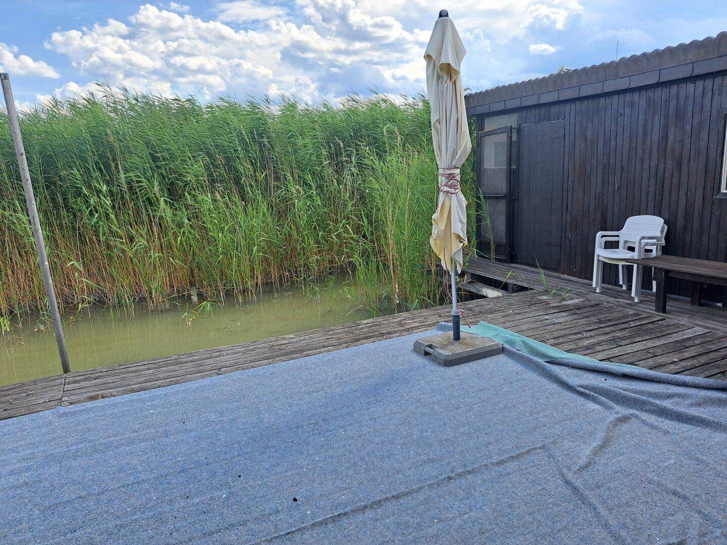 Terrassenbereich am Wasser mit einem geschlossenen Sonnenschirm und Blick auf das dichte Schilf.