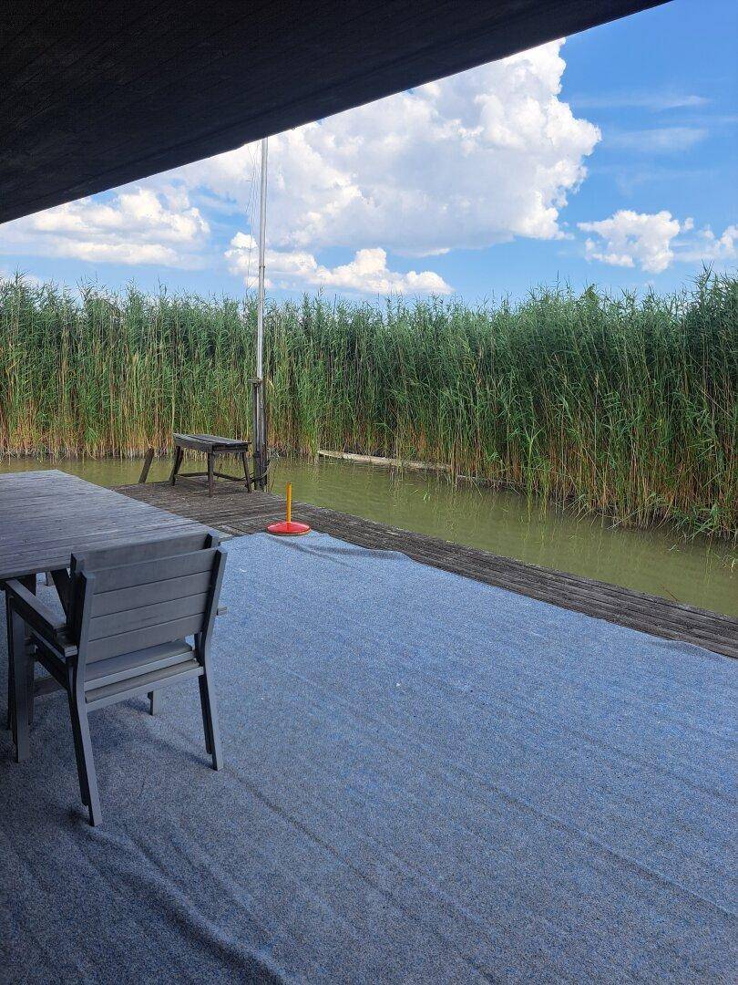 Möblierte Terrasse mit Esstisch und Stühlen, direkt am Wasser mit üppigem Schilf.